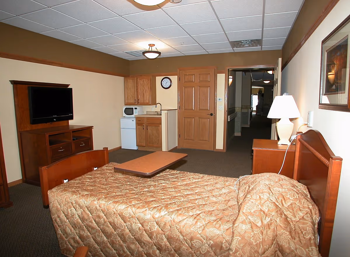 A senior living facility room with a single bed covered in a patterned beige bedspread, a wooden headboard, and a bedside table with a lamp. There is a wooden TV stand with a flat-screen TV mounted on the wall. A small kitchenette area with a microwave, mini fridge, sink, and wooden cabinets is visible. The room has beige walls with a brown trim and a ceiling with recessed lighting. A hallway is visible through an open door.