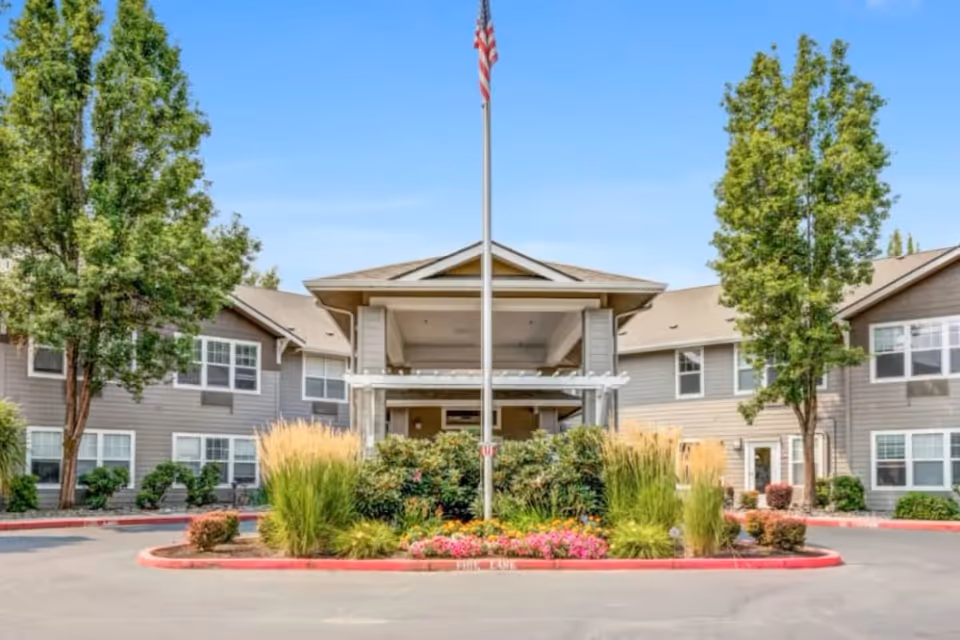 Front exterior view of Woodside Senior Living facility with a covered entrance, an American flag on a flagpole, landscaped bushes, flowers, and trees on a sunny day.