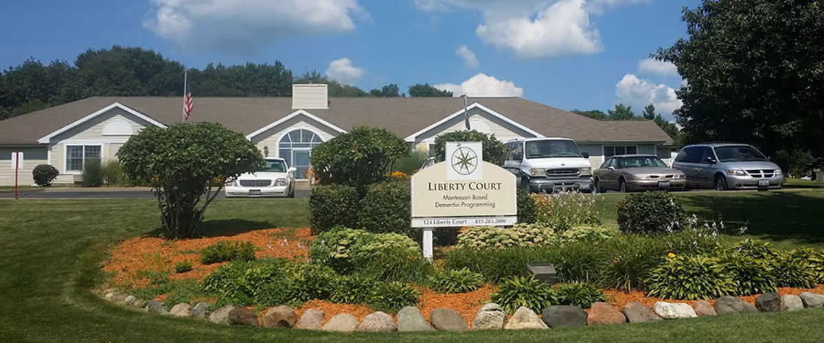 Exterior view of Liberty Court facility with a landscaped garden in the foreground featuring bushes, flowers, and mulch bordered by rocks. Several cars are parked in front of a single-story building with a beige exterior and a brown roof under a partly cloudy blue sky.