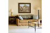 A cozy living room with a beige sofa adorned with two light blue pillows and a gray throw blanket. There is a glass coffee table with a metal frame in front of the sofa. On the left side of the sofa, there is a wooden side table with a lamp, and on the right side, a tall floor lamp. A framed painting of a scenic village hangs on the wall above the sofa.