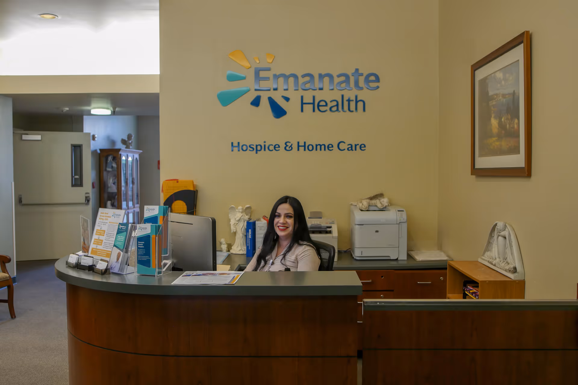 Reception area of Emanate Health Hospice & Home Care with a smiling receptionist sitting behind a curved wooden desk. The wall behind her displays the Emanate Health logo and the words Hospice & Home Care. The area includes office equipment, brochures, and framed artwork on the wall.