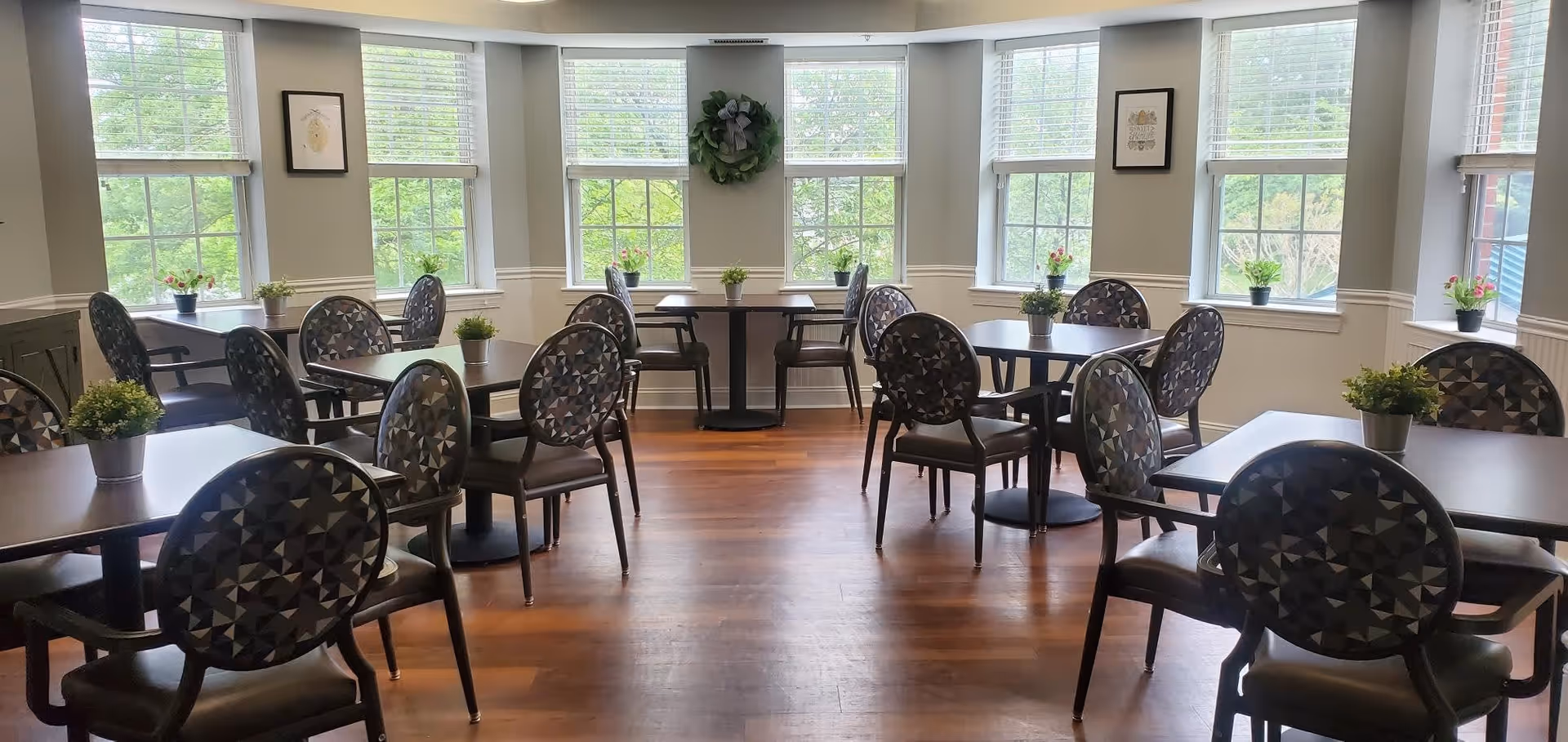 A bright dining room with multiple tables and chairs arranged neatly. Each table has a small potted plant as a centerpiece. Large windows with white blinds allow natural light to fill the room, and framed artwork and a wreath decorate the walls.