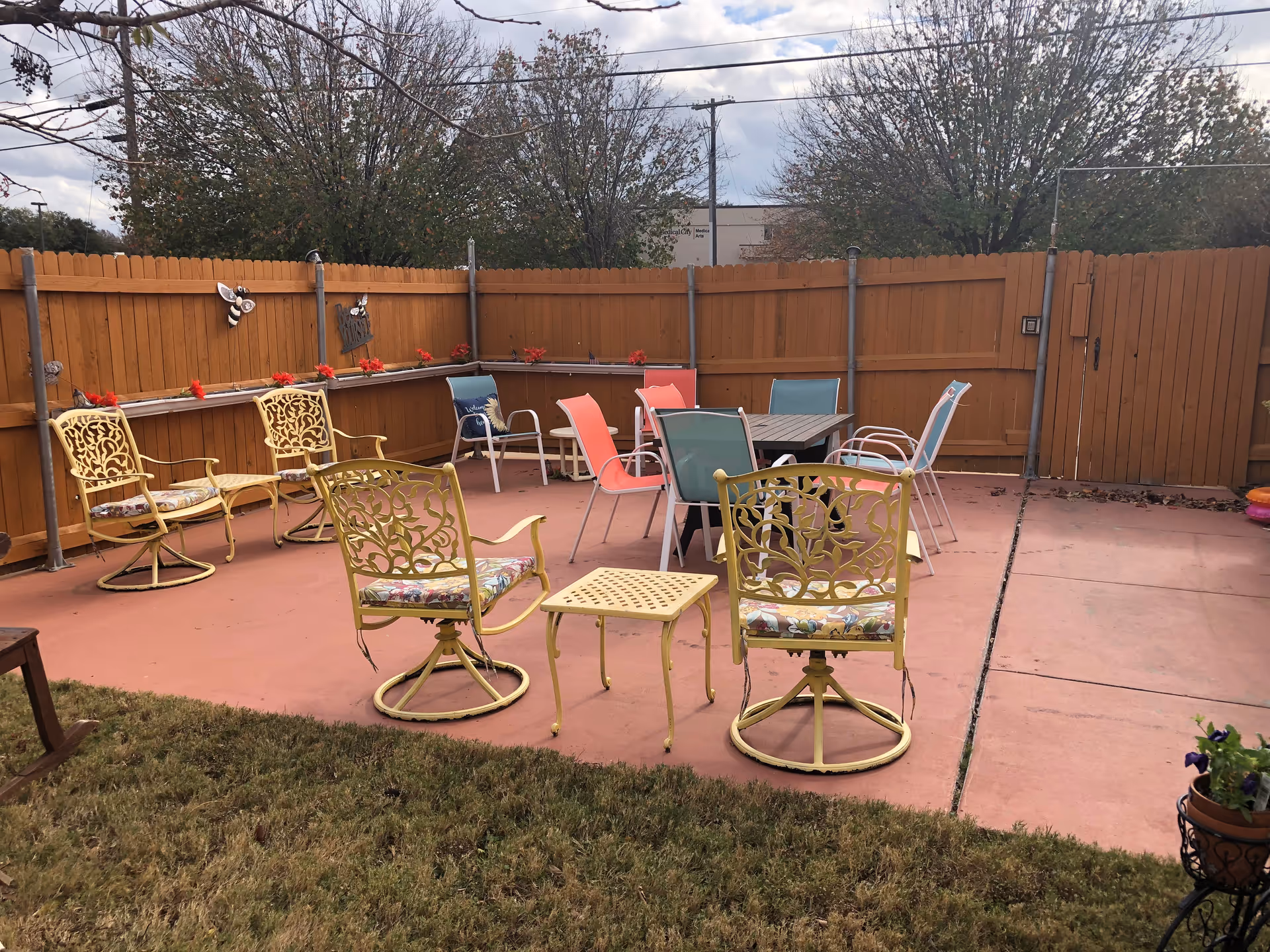 Outdoor patio area with a variety of chairs including yellow metal chairs with floral cushions, colorful plastic chairs, and a table in the center. The patio is enclosed by a wooden fence with some decorative items and red flowers along the fence. Trees and a cloudy sky are visible in the background.
