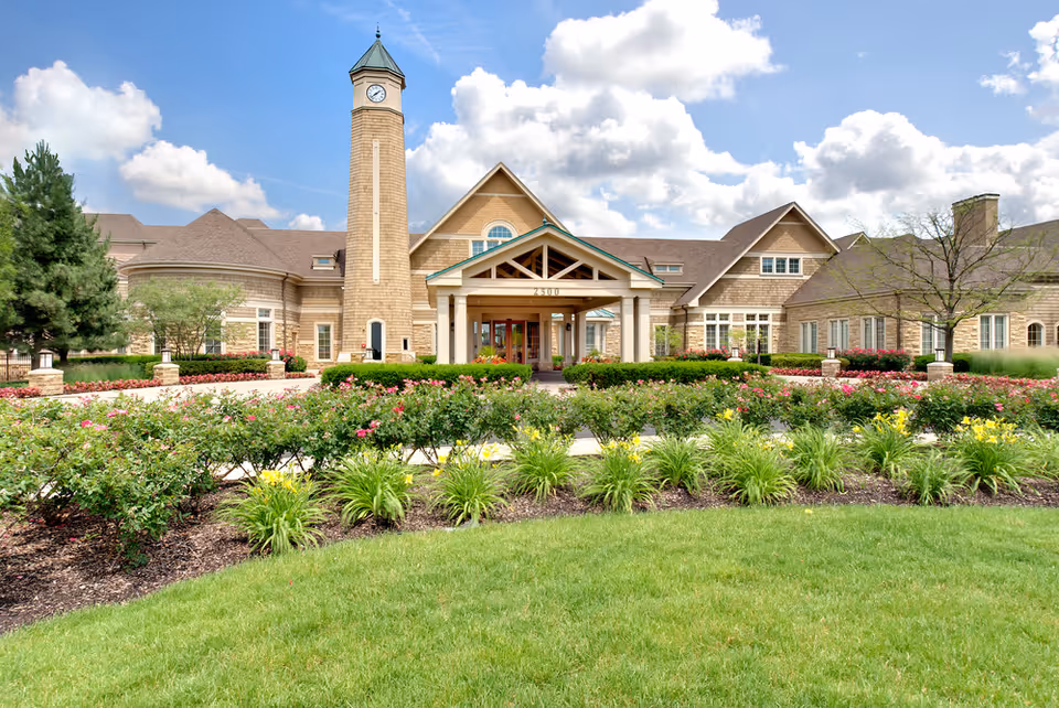 Front exterior view of a senior living facility named Vi at The Glen, featuring a large building with a clock tower, a covered entrance, well-maintained flower beds, shrubs, and a green lawn under a partly cloudy sky.