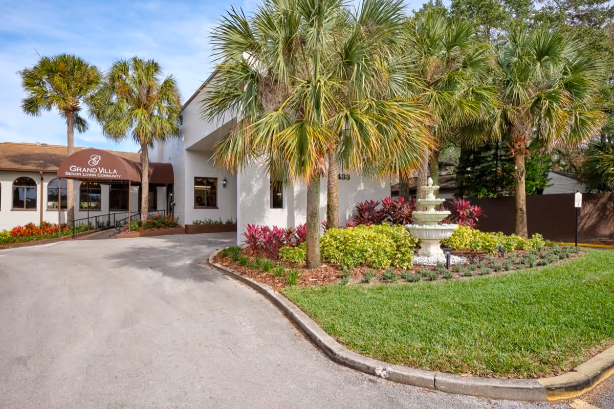 Exterior front entrance of the Grand Villa senior living community with a driveway, palm trees, landscaped fountain, and an awning over the entry.