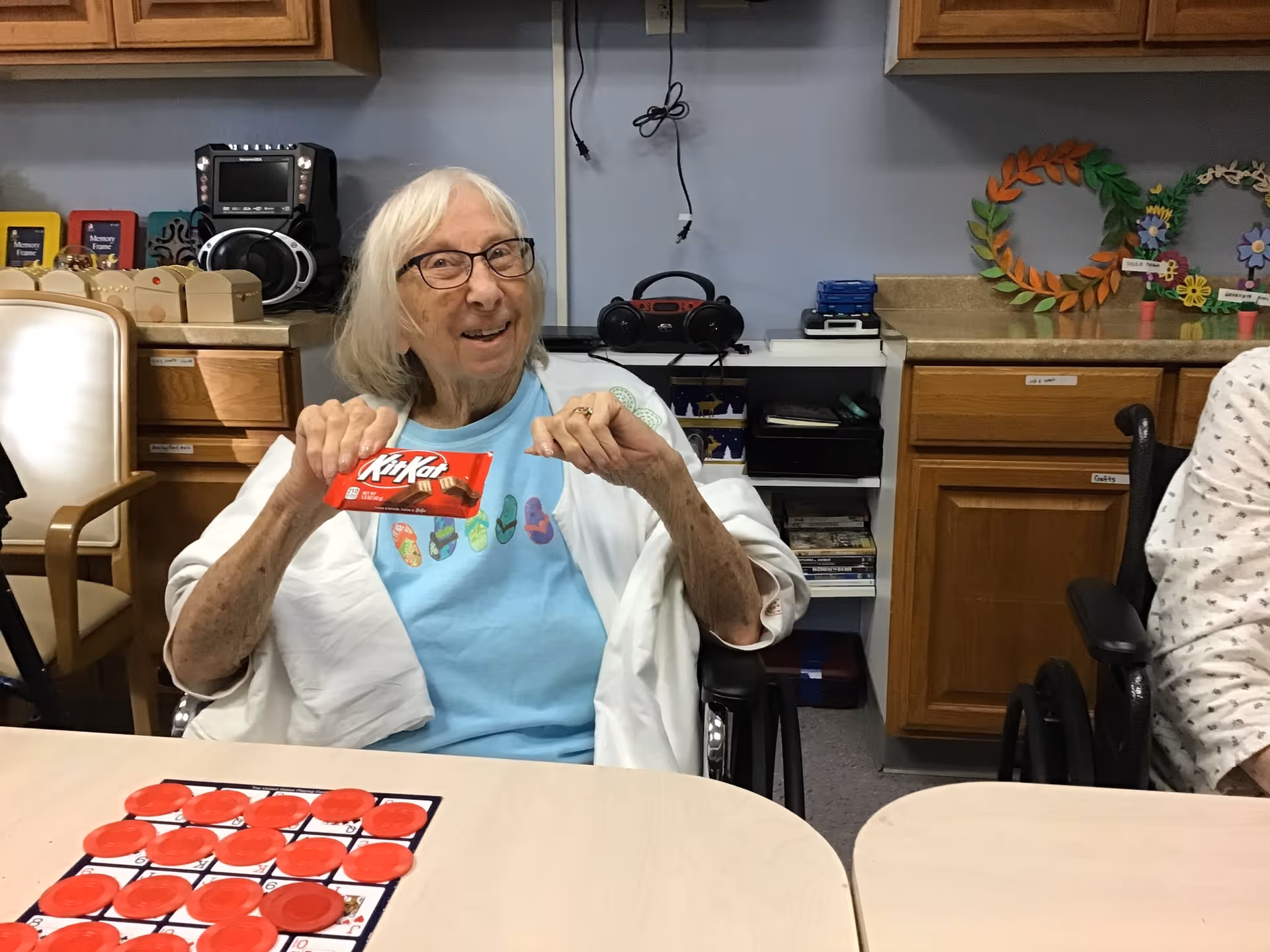 An elderly woman with white hair and glasses sitting in a wheelchair at a table, smiling and holding a KitKat chocolate bar. The table has a game board with red circular pieces. The background shows wooden cabinets, a radio, and colorful wreath decorations on the counter.