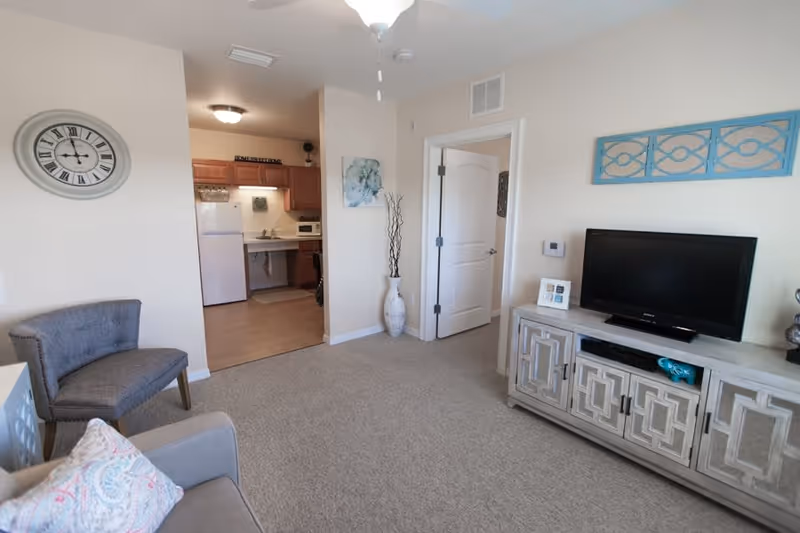 A cozy living room area with a gray armchair and a sofa with a patterned pillow. A wall clock hangs on the left wall. The room opens into a small kitchen with wooden cabinets, a white refrigerator, and a microwave. On the right side, there is a TV on a decorative cabinet with a blue wall art piece above it. A white door and a tall vase with decorative branches are also visible.