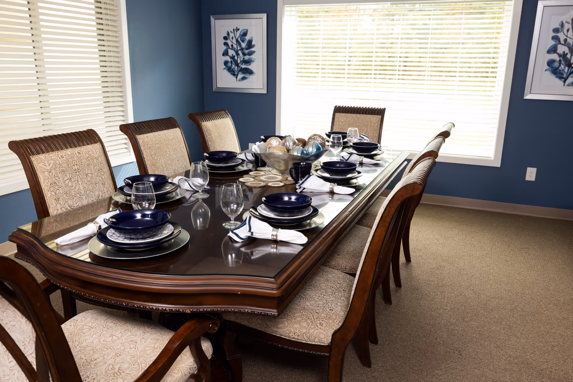 A formal dining room with a long wooden table set for eight people. The table has a glass top and is arranged with blue plates, bowls, glasses, and white napkins. The room has blue walls, beige carpet, two windows with white blinds, and framed botanical artwork on the walls.