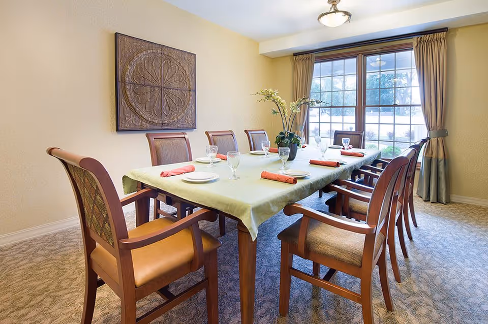 A dining room with a long rectangular table covered with a light green tablecloth, set with plates, glasses, and folded red napkins. There are eight wooden chairs with cushioned seats around the table. A decorative wall art piece hangs on the left wall, and a large window with beige curtains lets in natural light.