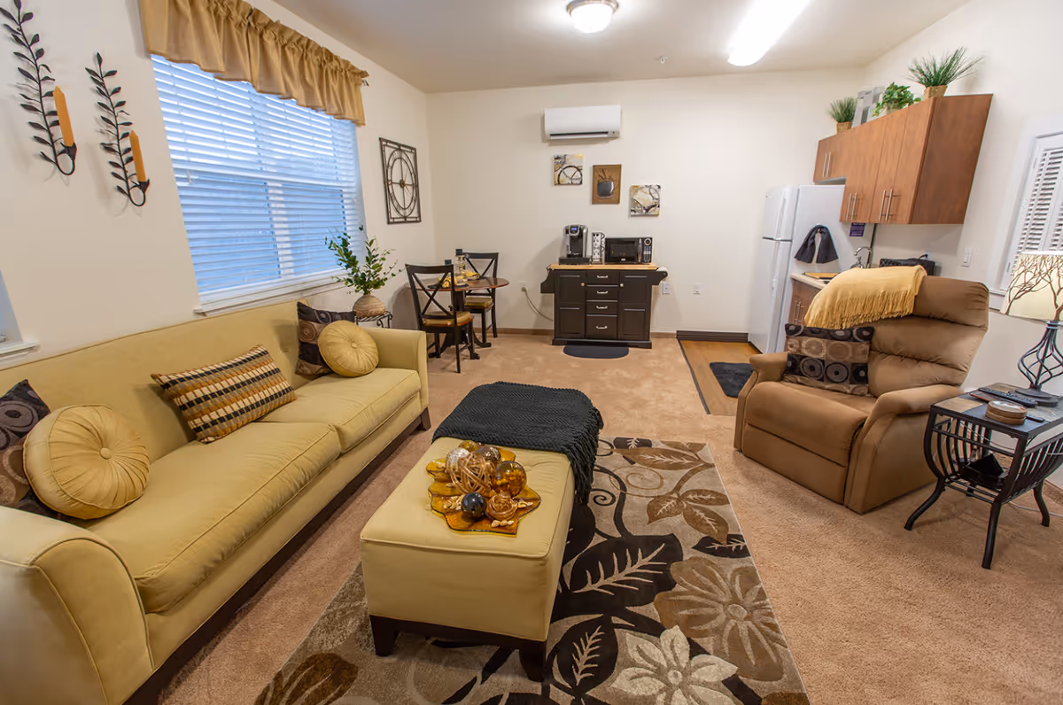 Cozy living room with a yellow sofa, ottoman, recliner, small dining table and kitchenette in the background.
