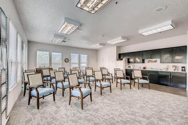 A bright room with multiple rows of wooden chairs with light blue cushions arranged facing the front. The room has large windows on one side letting in natural light. At the front, there is a kitchenette area with black cabinets, a microwave, coffee maker, and decorative signs on the wall. The ceiling has several rectangular fluorescent light fixtures.