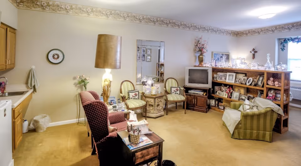 A cozy living room area in a senior living facility with beige walls and carpet. The room features a small kitchenette with wooden cabinets on the left, a vintage-style lamp, two green striped chairs, a wooden bookshelf filled with framed photos and decorative items, a small TV on a wooden stand, and a green armchair with a white blanket. There is a window with floral curtains and an air conditioning unit beneath it.