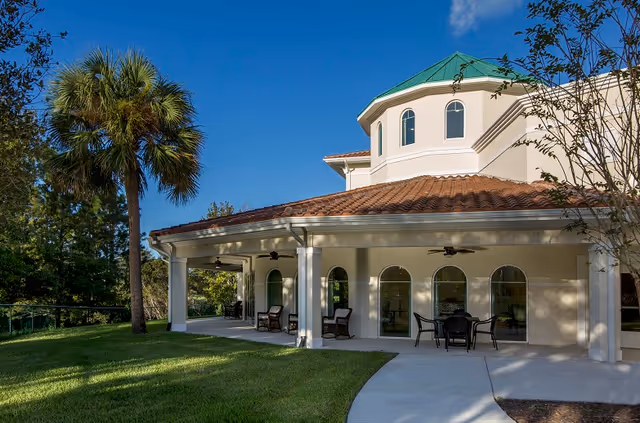 Exterior view of a senior living facility with a covered patio area featuring outdoor seating and ceiling fans. The building has a tiled roof and arched windows, surrounded by green grass, trees, and a clear blue sky.