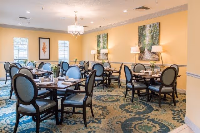 A dining room with several dark wood tables and blue cushioned chairs arranged neatly. The room has yellow walls adorned with framed artwork and two windows letting in natural light. The floor is covered with a patterned carpet in shades of blue and beige. There are table settings with cups, plates, and small flower arrangements on each table.