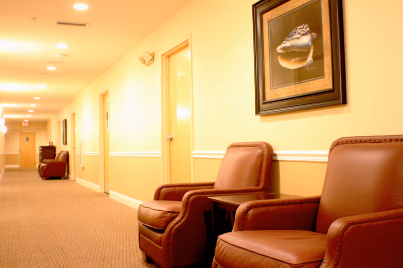 A well-lit hallway in an assisted living facility with beige walls and carpeted floor. There are several closed doors along the hallway, and two brown leather armchairs with a small table between them are positioned against the wall. A framed picture of a seashell hangs above the chairs.