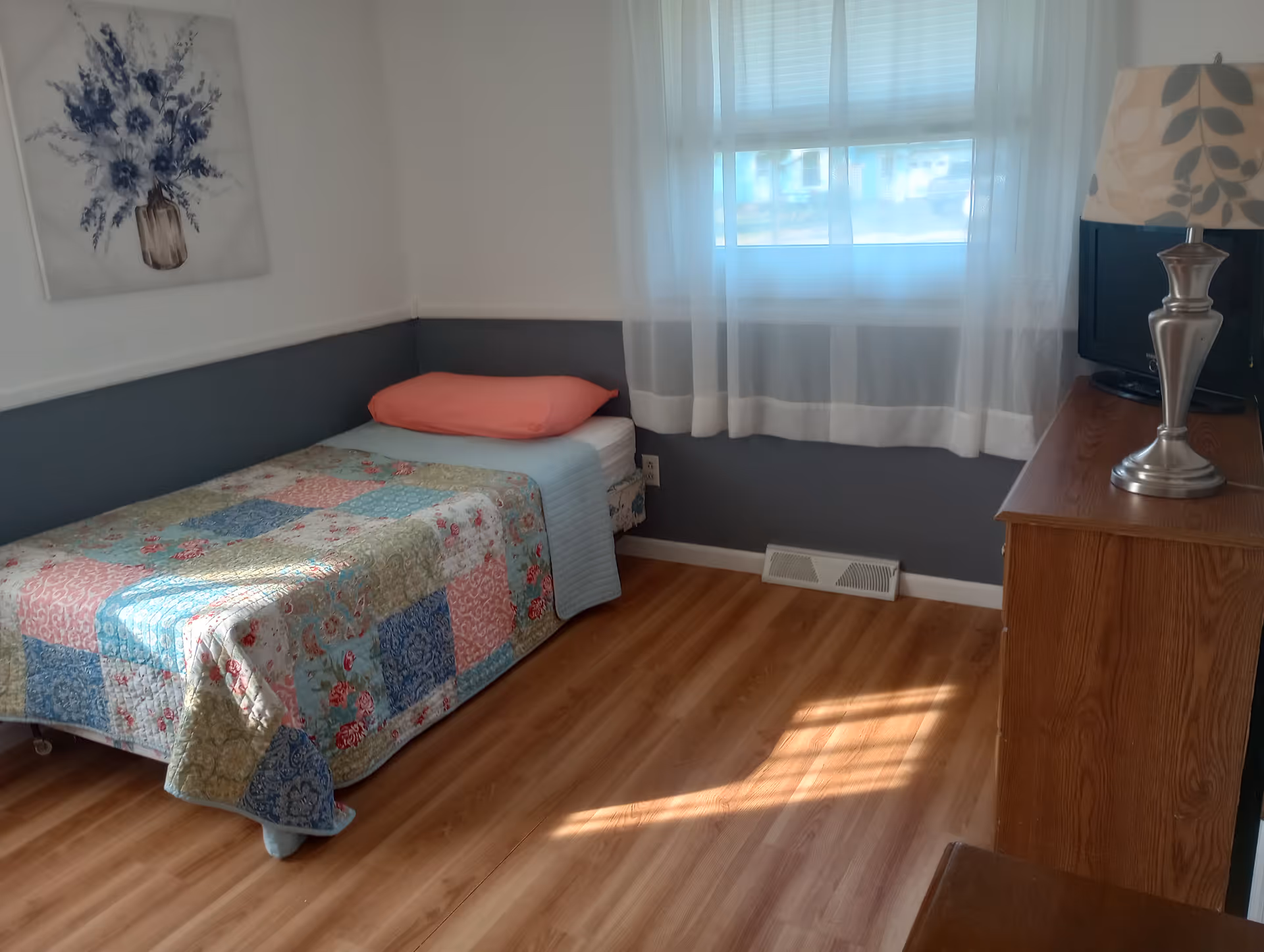 A small, cozy bedroom with a single bed covered in a colorful patchwork quilt and a coral pillow. There is a wooden dresser with a silver lamp and a small TV on top. A window with sheer white curtains lets in natural light, and a floral painting hangs on the wall above the bed. The room has wood flooring and a two-tone wall with white on top and gray on the bottom.