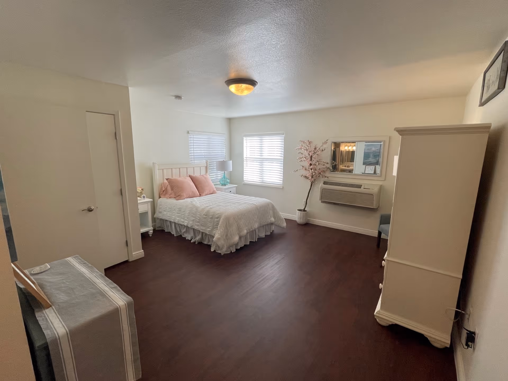 A bright and spacious bedroom with a white bed frame, white bedding, and two pink pillows. The room has dark wooden floors, two windows with white blinds, a white nightstand with a lamp, a white armoire, a small table covered with a cloth, a potted plant, and a wall-mounted air conditioning unit beneath a mirror.