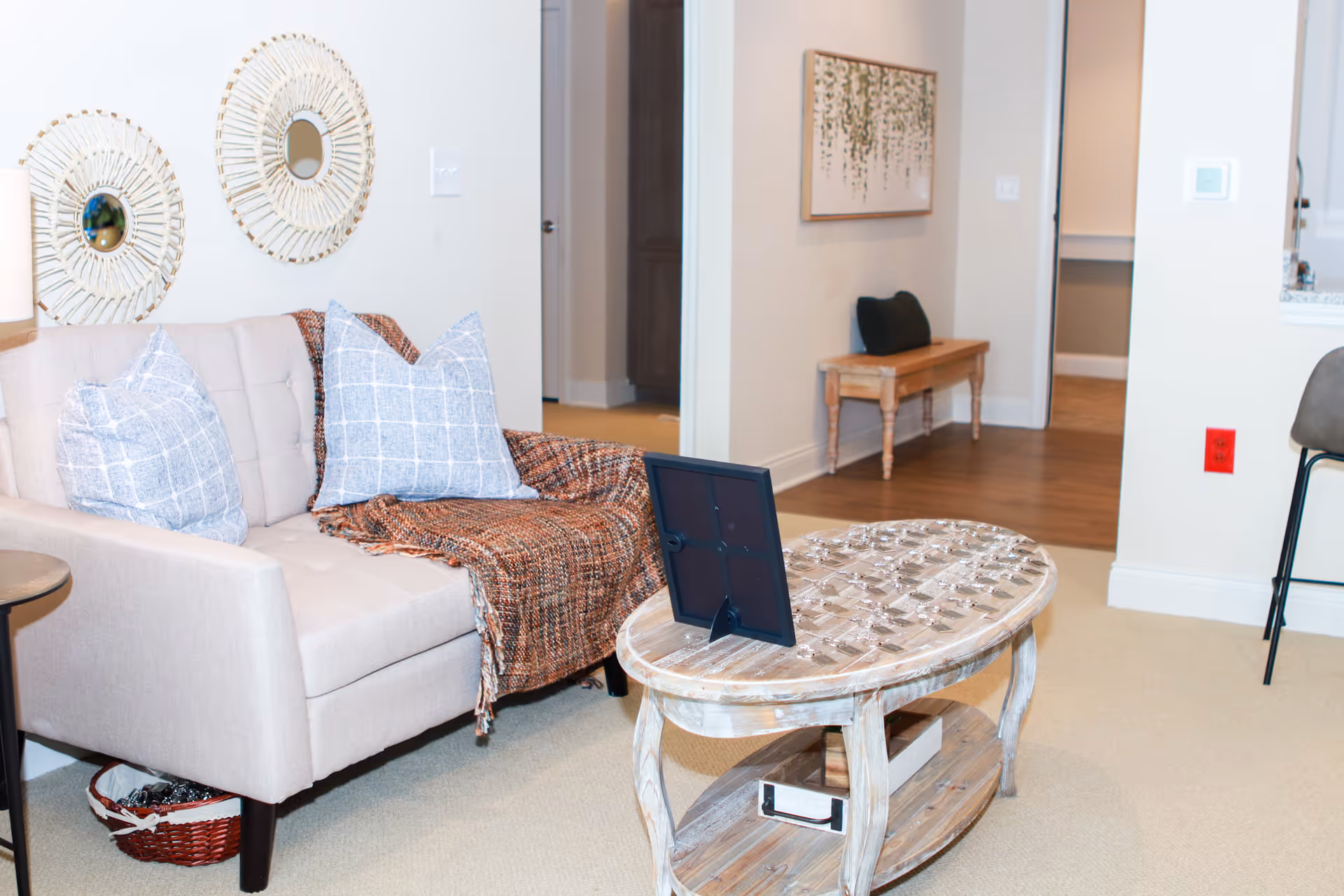 A cozy living room area with a beige loveseat adorned with two blue checkered pillows and a brown throw blanket. A rustic wooden coffee table with a framed photo sits in front of the loveseat. Two decorative round mirrors hang on the wall above the loveseat. In the background, a wooden bench with a dark cushion is placed against the wall, and doorways lead to other rooms. The floor is carpeted near the loveseat and transitions to wood flooring in the hallway.