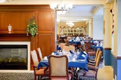 Dining room with multiple tables set with blue tablecloths, upholstered chairs, chandeliers, and a fireplace.