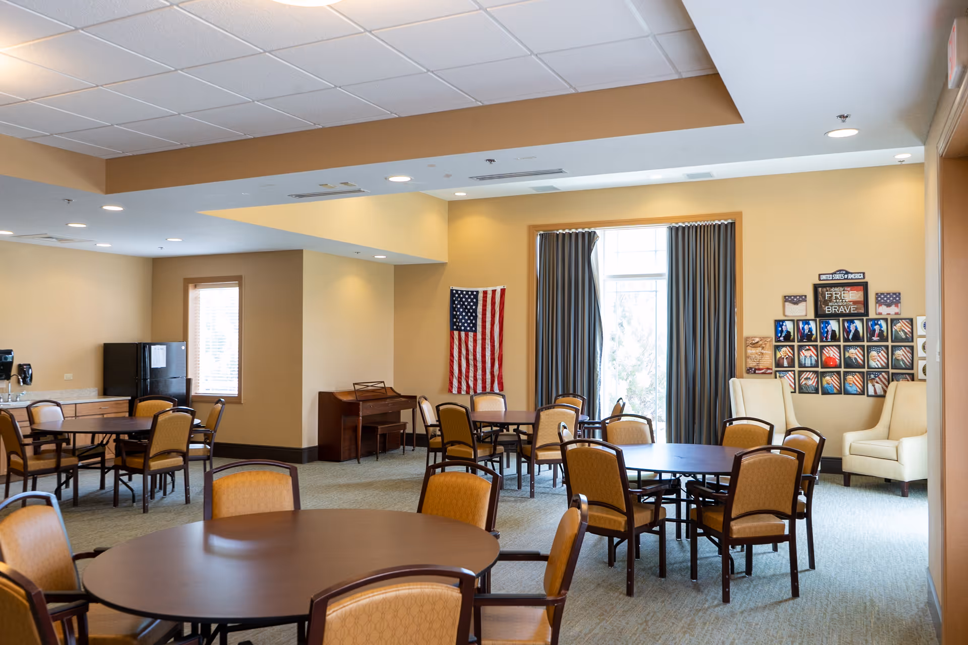 A spacious room with multiple round tables and chairs arranged for group seating. There is a piano against the far wall, an American flag hanging on the wall, and a large window with curtains letting in natural light. The walls are decorated with patriotic-themed pictures and plaques. The room has a warm, inviting atmosphere with beige walls and carpeted floor.