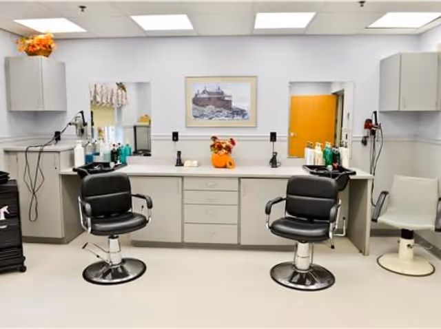Interior view of a hair salon area with two black salon chairs and one white chair in front of a long counter with mirrors and various hair care products. The room has white walls, cabinets, and bright overhead lighting. A framed picture hangs on the wall above the counter.