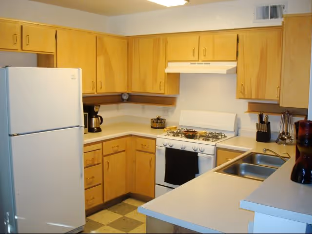 A kitchen with light wood cabinets, a white refrigerator, a white gas stove with a black towel hanging on the handle, a coffee maker on the counter, a double sink, and various kitchen utensils on the countertop.