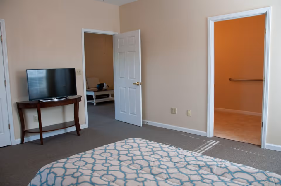 View of a bedroom in a senior living facility with a bed covered in a patterned comforter in the foreground. A flat-screen TV is placed on a wooden console table against the wall. Two open doorways lead to other rooms, one showing a glimpse of a sitting area with a chair and coffee table, and the other showing a room with a handrail on the wall.