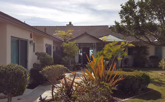 Exterior view of a single-story assisted living facility with a tiled roof, surrounded by various shrubs, small trees, and landscaped garden beds. A paved walkway leads to the entrance of the building under a small covered porch.