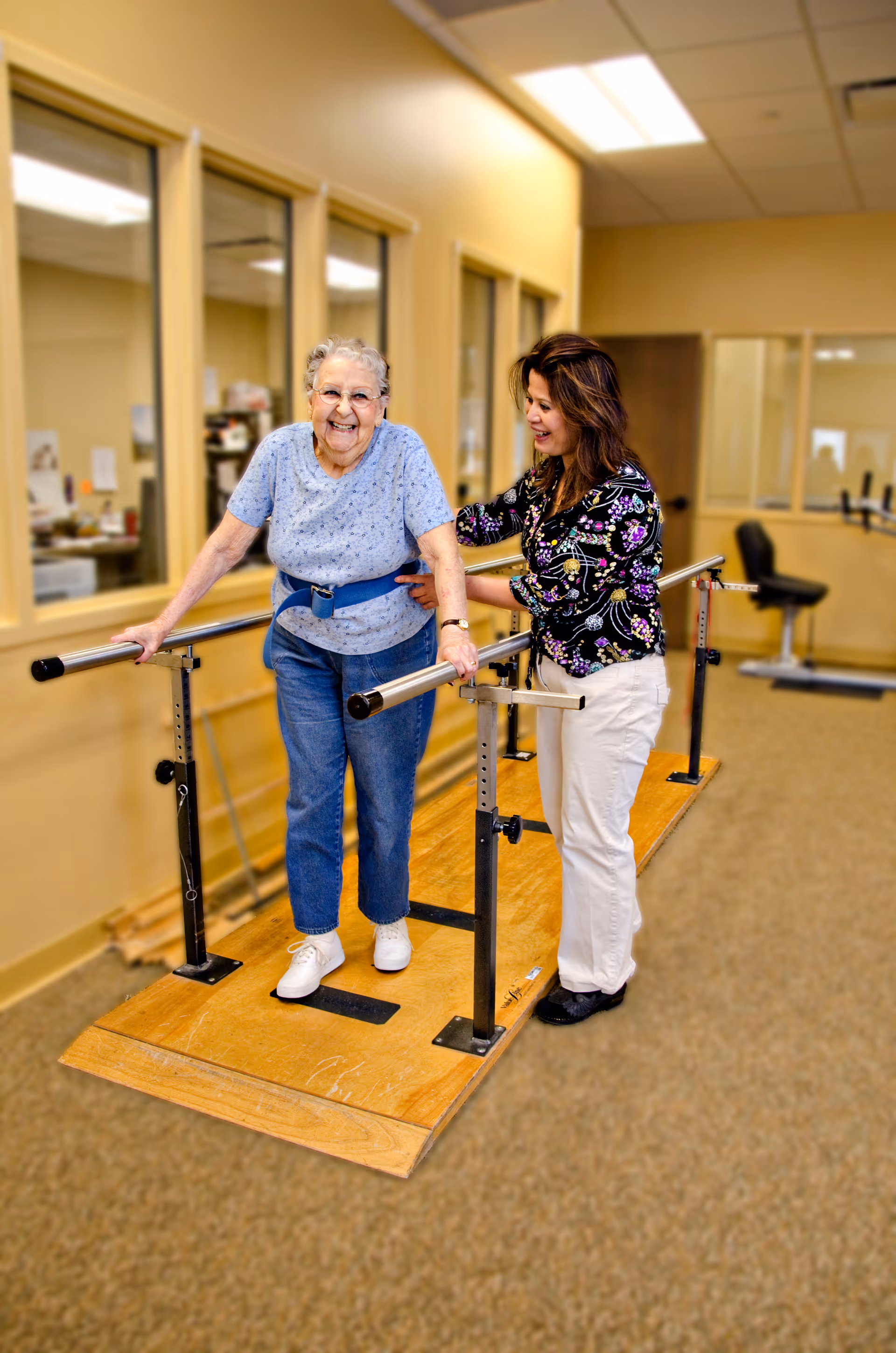 An elderly woman using parallel bars for walking exercise is assisted by a caregiver in a rehabilitation or therapy room with beige walls and carpeted floor.