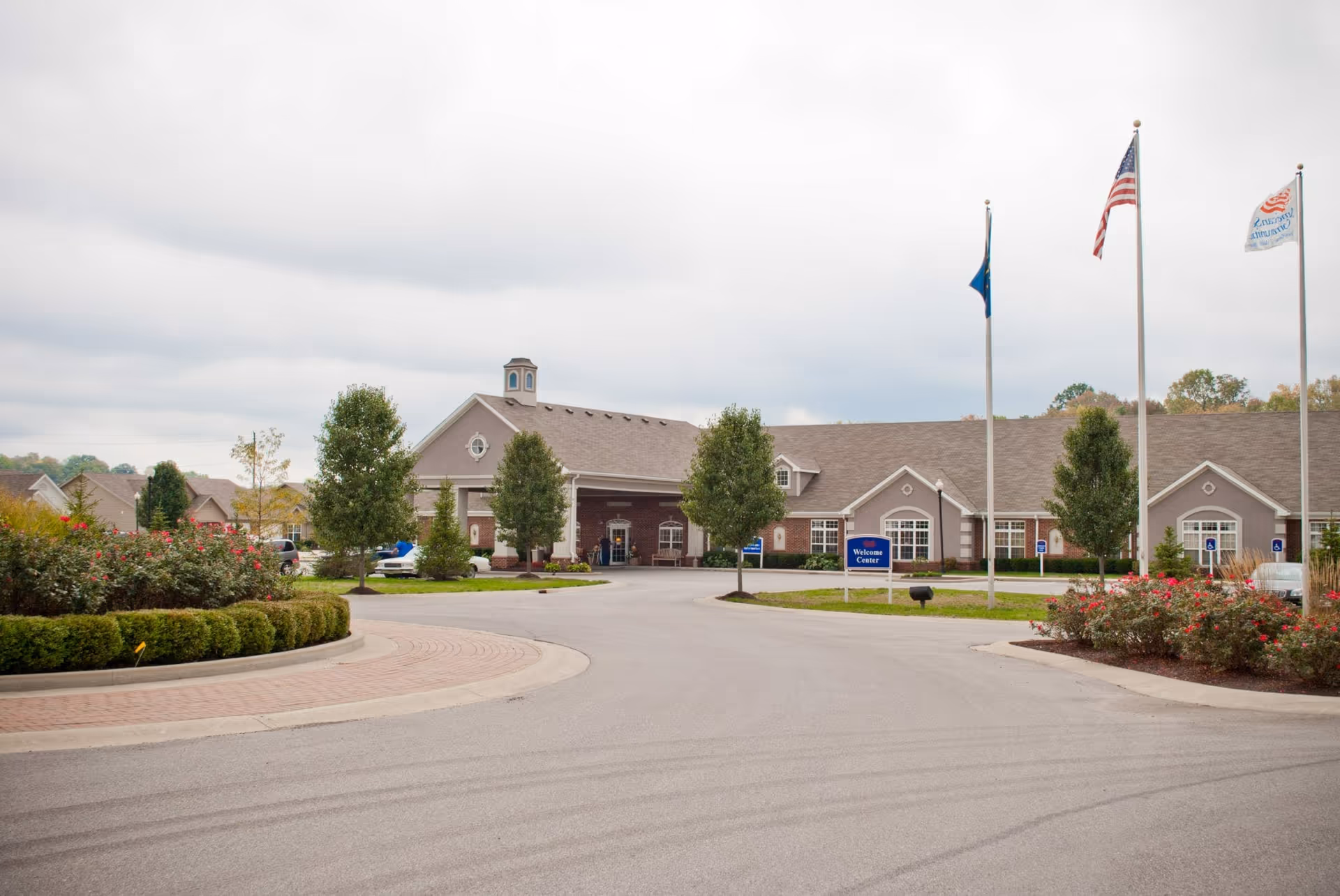 Exterior view of Meadow Lakes facility showing a large building with a peaked roof, a driveway leading to the entrance, landscaped bushes and trees, and three flagpoles with flags including the American flag. A blue sign near the entrance reads 'Welcome Center'.