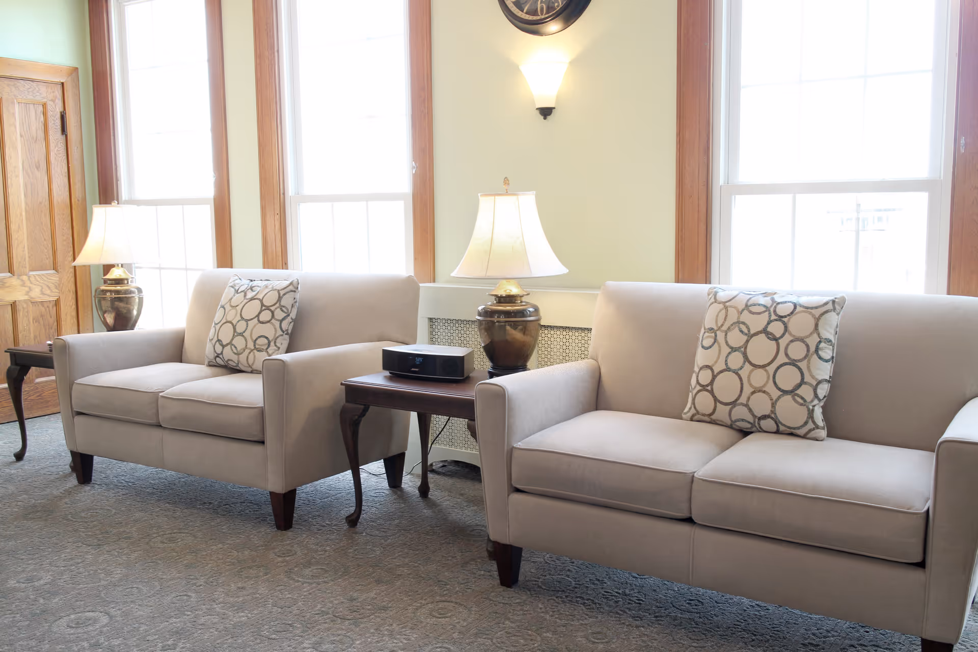 A bright living room area with two beige sofas, each with a decorative pillow featuring a circular pattern. Between the sofas is a wooden side table with a lamp and a small electronic device. Large windows with wooden frames allow natural light to fill the room, and a wall-mounted clock and sconce light are visible above the sofas.