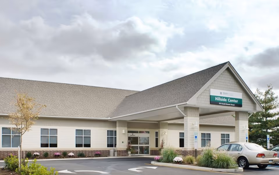 Exterior view of a single-story building with a covered entrance labeled Hillside Center. There are several windows along the building, a few small trees and shrubs in front, and a parked car near the entrance. The sky is cloudy.