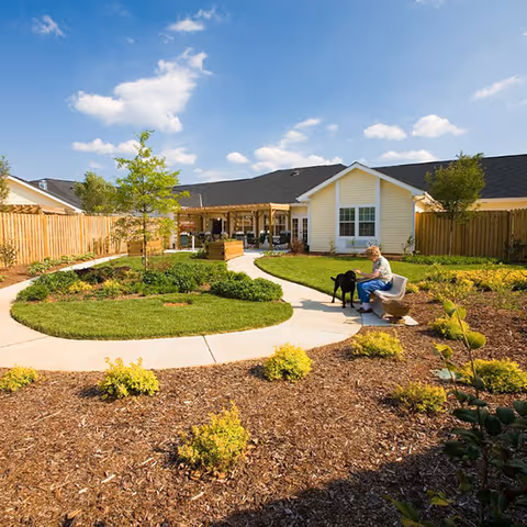 A sunny outdoor garden area at a senior living facility with a curved concrete pathway, green grass, small shrubs, and a wooden fence. A senior woman is sitting on a bench petting a black dog. The building in the background is light yellow with white trim and has a covered patio area with seating.