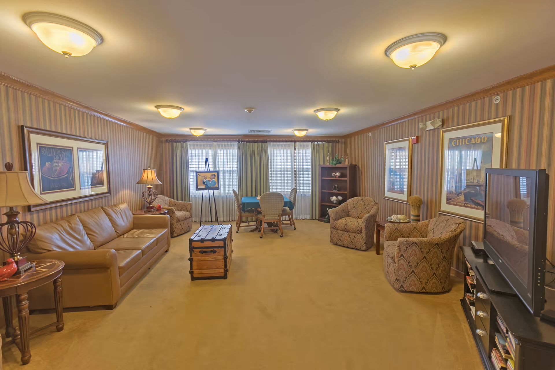 A cozy living room in a senior living facility with a tan leather sofa, two patterned armchairs, a wooden trunk coffee table, a TV on a stand, framed artwork on striped wallpapered walls, and a dining table with chairs near large windows with curtains.