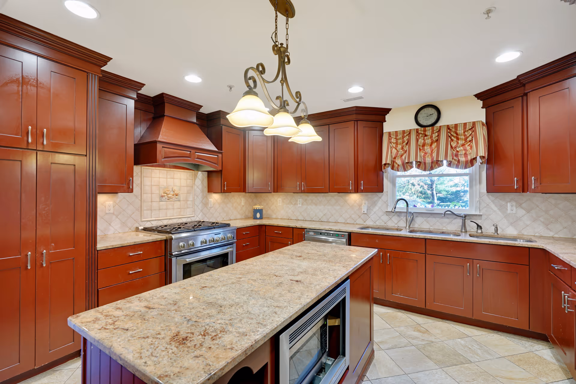 A spacious kitchen with dark wood cabinets, a large island with a granite countertop, a stainless steel stove and oven, a dishwasher, and a double sink under a window with a striped valance. The kitchen has tiled floors and recessed lighting with a decorative hanging light fixture above the island.