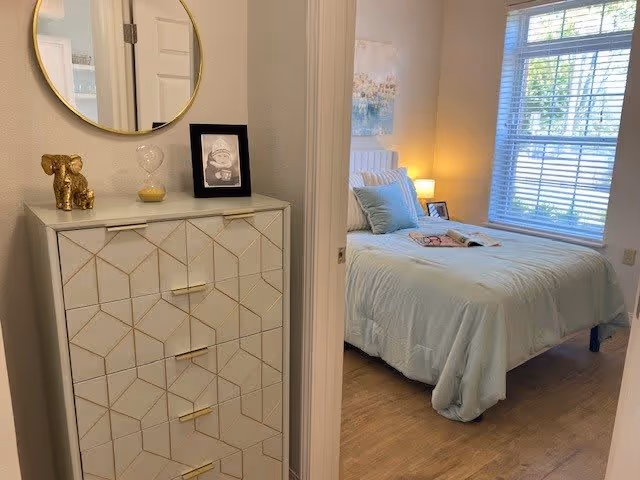 View into a bedroom from a hallway showing a bed with light blue bedding and pillows, a bedside table with a lamp, a window with blinds, and a decorative chest of drawers with a round mirror, a gold elephant figurine, an hourglass, and a framed picture on top.