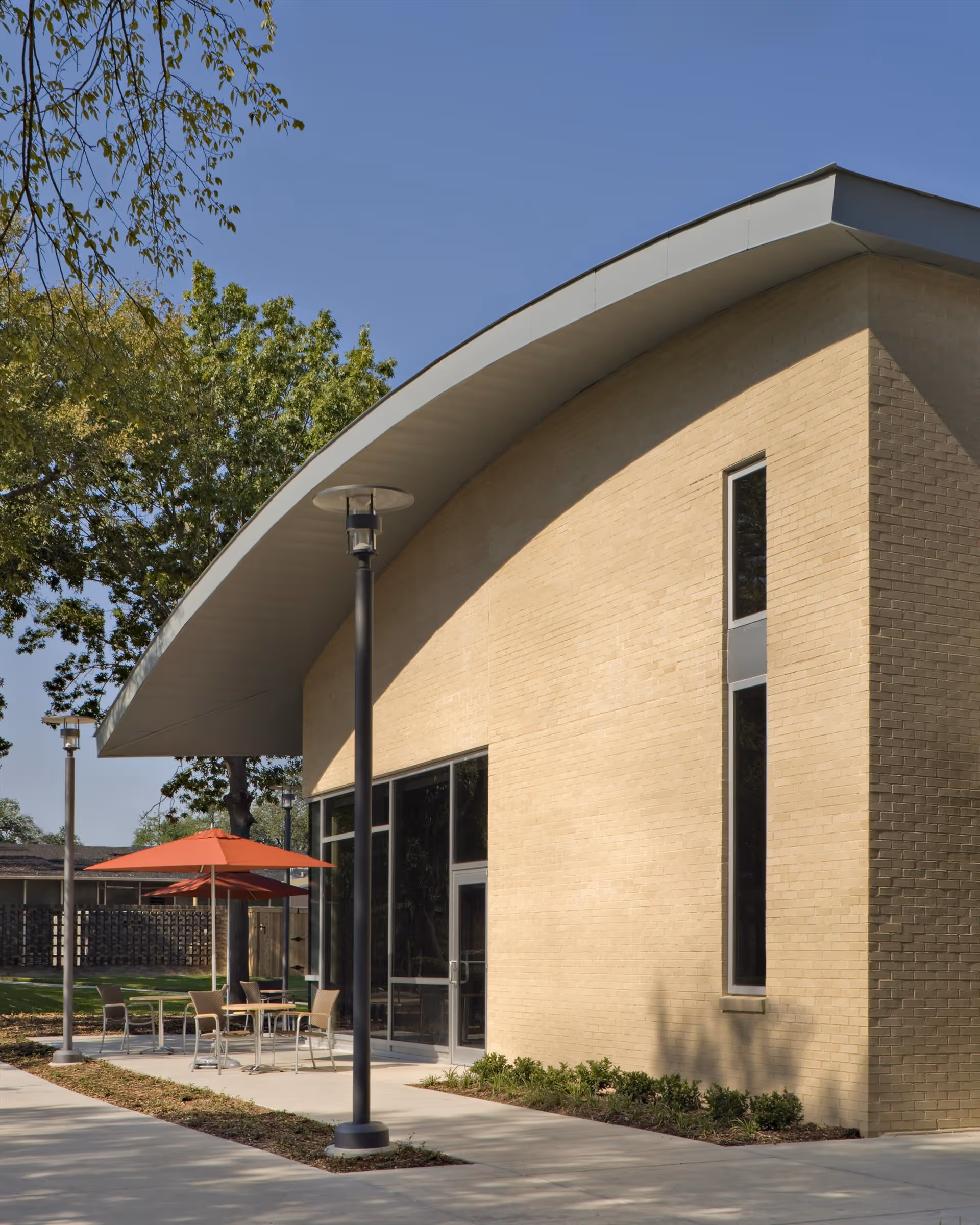 Exterior view of a modern building with light-colored brick walls and a curved roof. There is a patio area with tables, chairs, and red umbrellas, surrounded by greenery and trees under a clear blue sky.