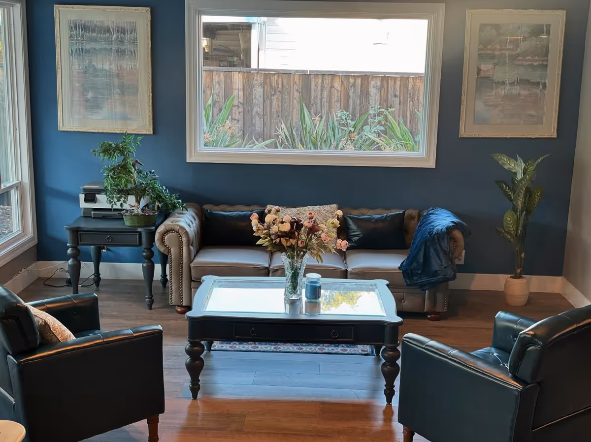 Cozy living room with a tufted leather sofa and two armchairs facing a glass-top coffee table topped with a vase of flowers, set against a blue wall with a large window.