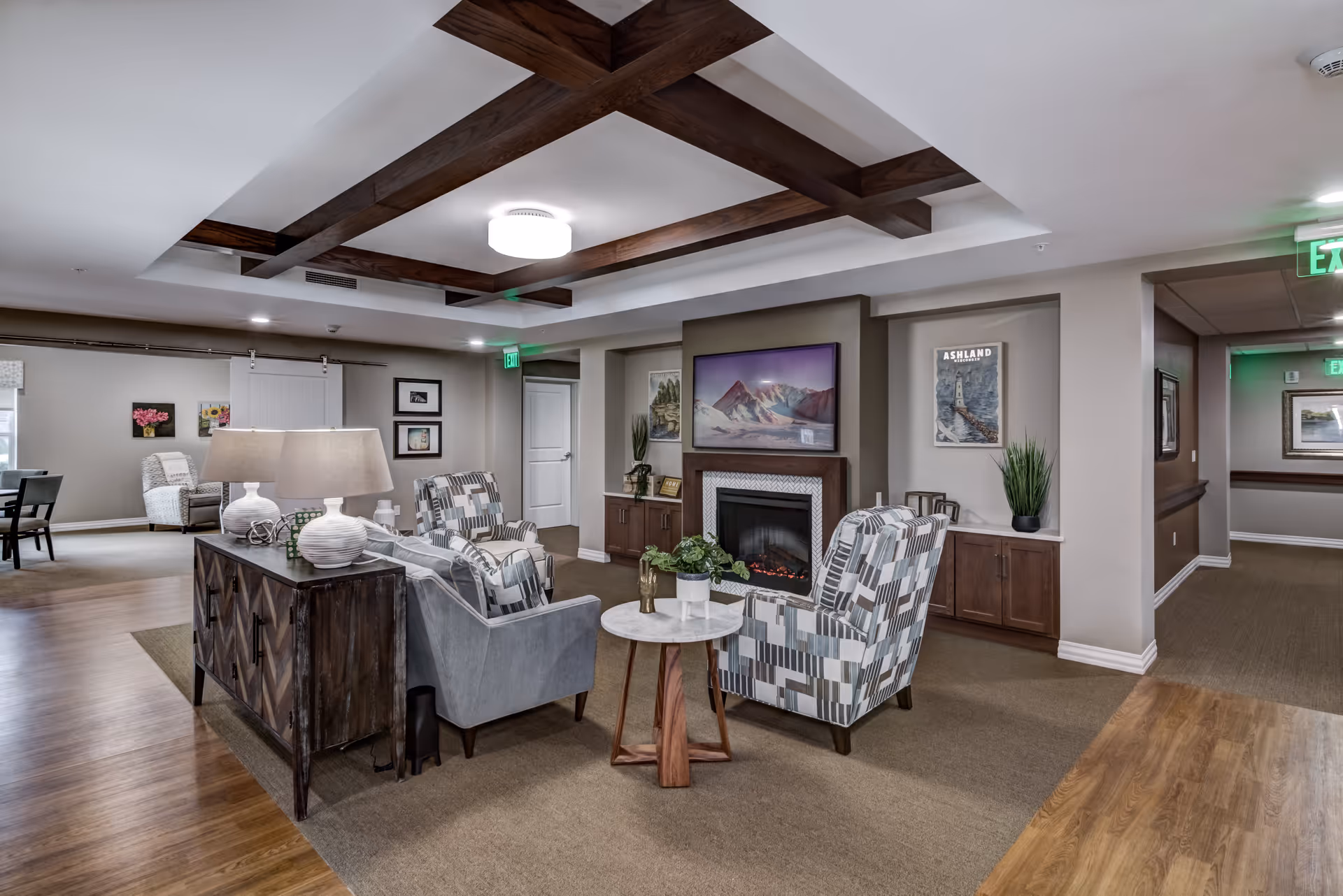 A cozy living room area in an assisted living facility featuring a fireplace with a mountain landscape painting above it. The room has a ceiling with wooden beams, two patterned armchairs, a gray sofa, a small round table with a plant, and a wooden console table with two lamps. The walls are decorated with framed pictures and there are exit signs visible in the background.