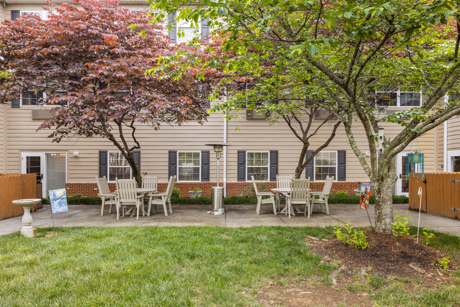 Outdoor courtyard patio with two dining tables and chairs under trees in front of a beige multiunit building.