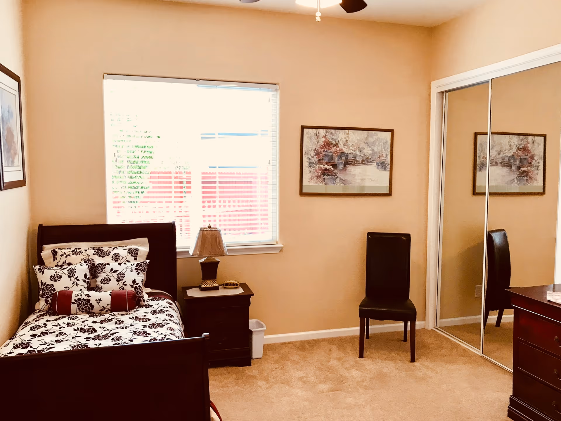 A cozy bedroom with a single bed featuring floral bedding, a wooden nightstand with a lamp, a black chair, a framed painting on the wall, a window with blinds, and a mirrored sliding closet door.