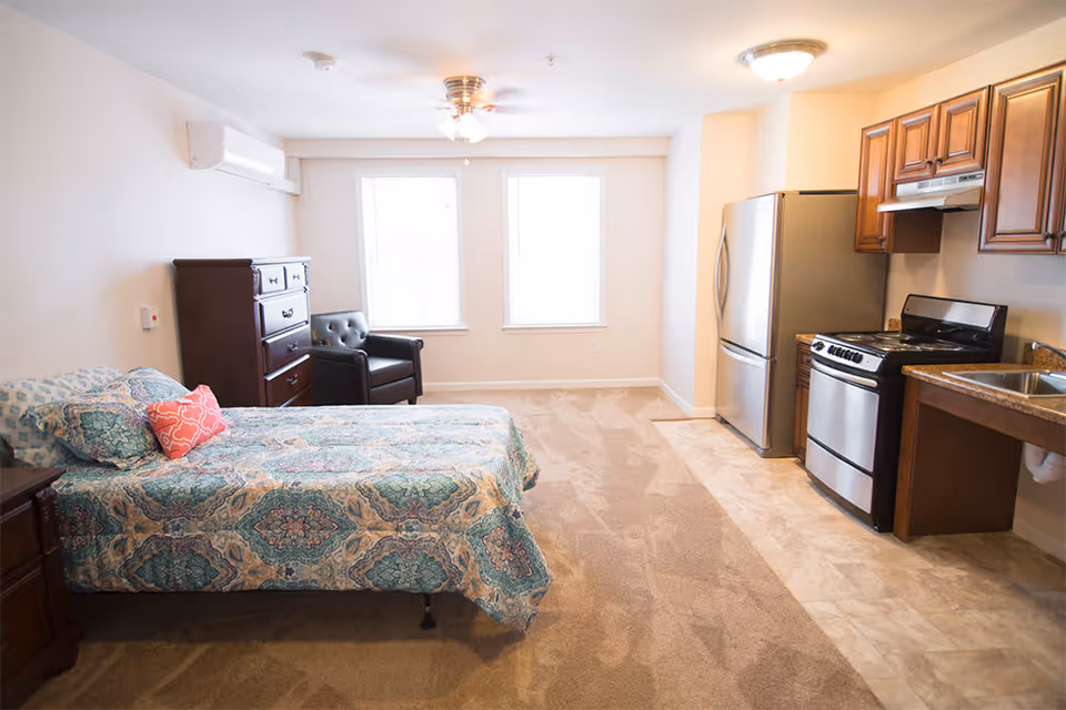 A bright and clean studio apartment featuring a bed with patterned bedding and pillows, a dark wooden dresser, a black armchair, two large windows, and a kitchenette with a stainless steel refrigerator, stove, and sink. The room has beige carpet and tile flooring, white walls, and ceiling lights including a ceiling fan.
