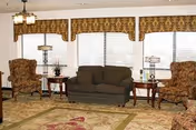 A living room area with a dark green sofa centered between two patterned armchairs. There are wooden side tables with lamps on either side of the sofa. The room has large windows with white blinds and patterned valances. A patterned area rug covers part of the floor, and a ceiling light fixture is visible.