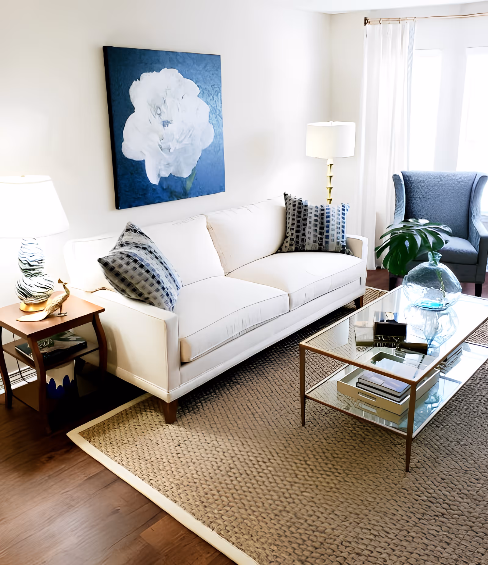 A bright living room with a white sofa adorned with patterned pillows, a glass coffee table with decorative items, a blue armchair near a window with white curtains, a side table with a lamp and a small sculpture, and a large painting of a white flower on a blue background hanging on the wall.