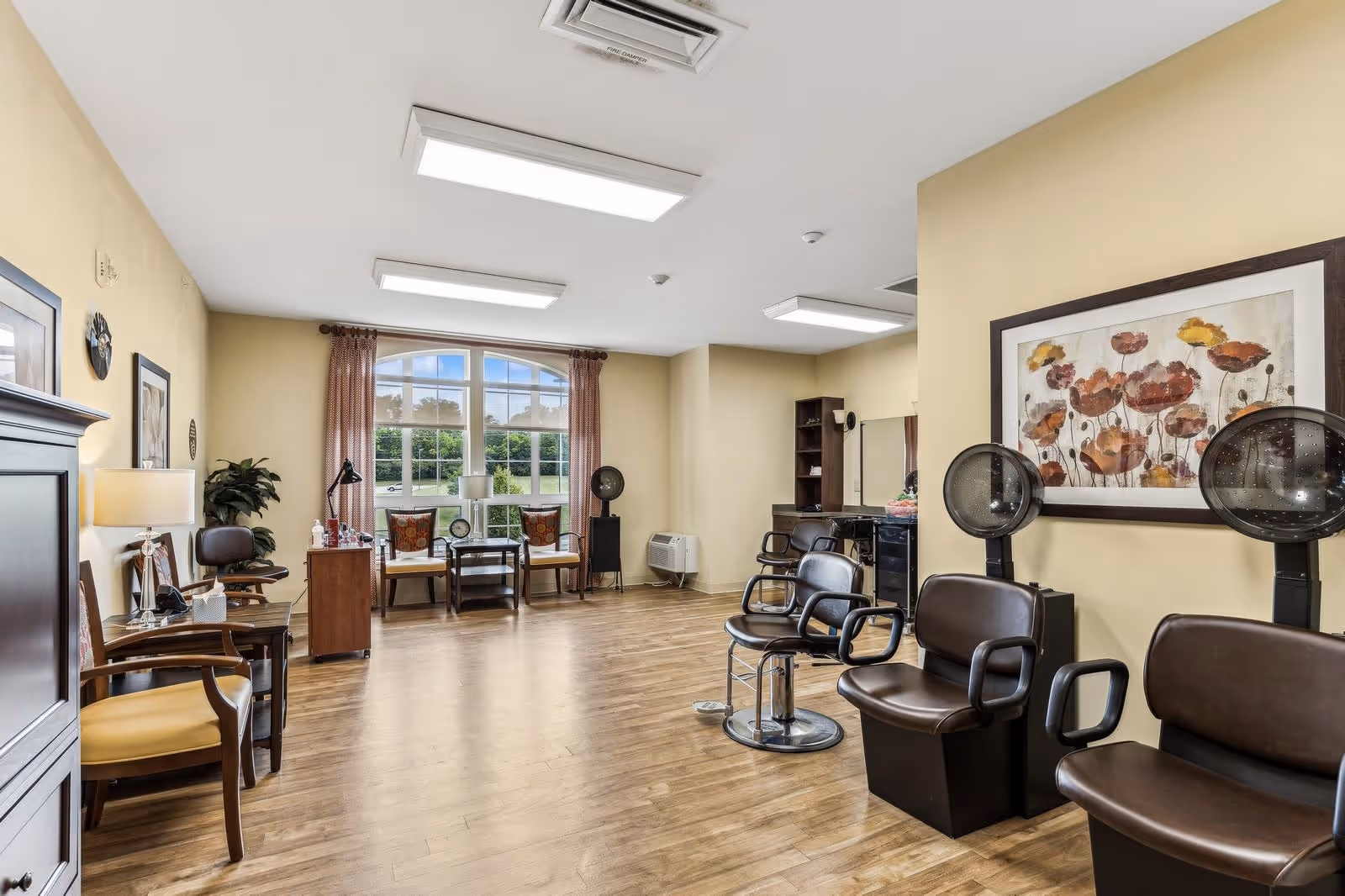 Interior of a salon room in a senior living facility with wooden flooring, three brown salon chairs with hair dryers, a large window with curtains, two chairs and a small table near the window, a plant, framed artwork on the walls, and bright ceiling lights.