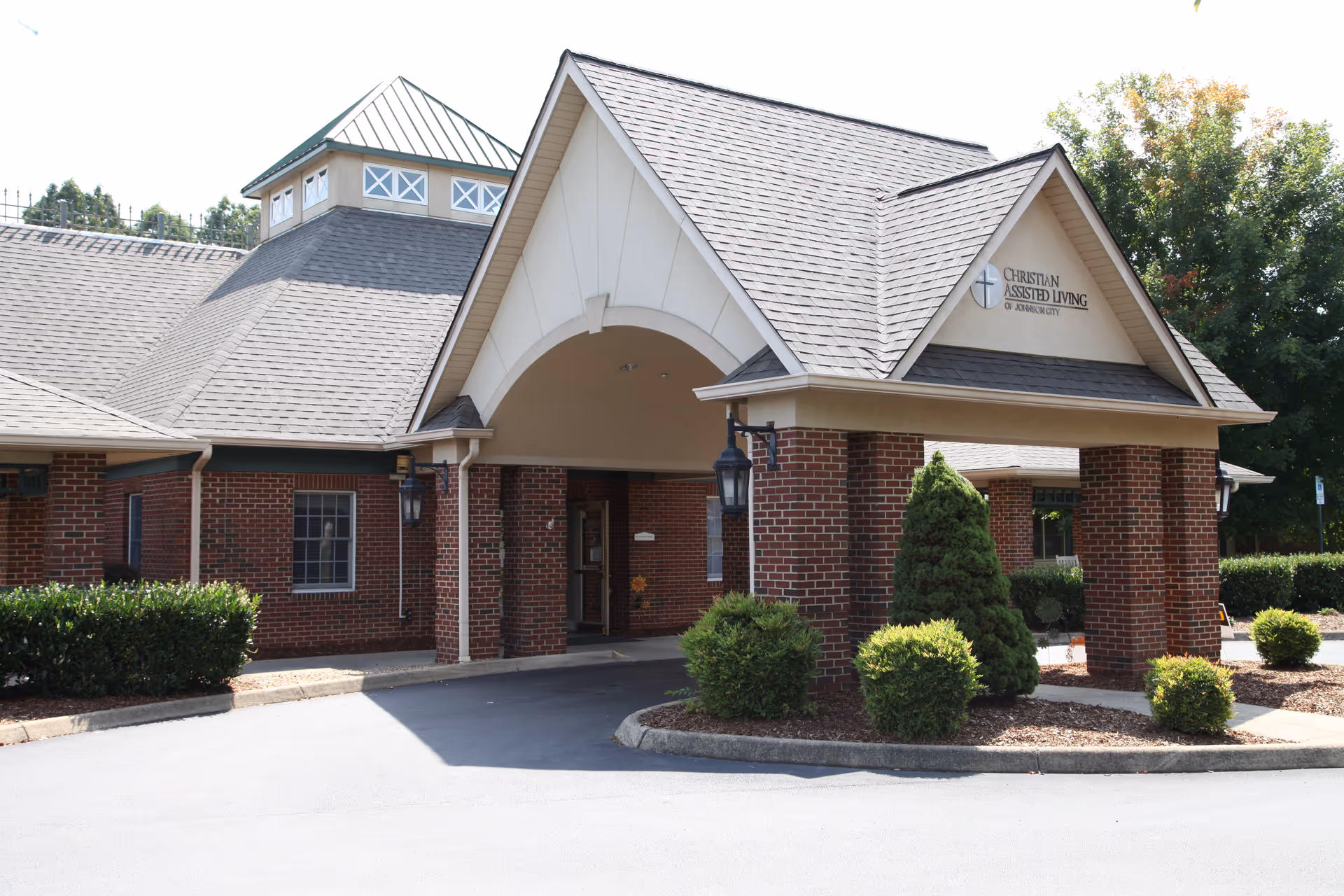 Exterior view of Christian Assisted Living of Johnson City building with a covered entrance, brick walls, and a peaked roof. There are bushes and small trees around the entrance area.