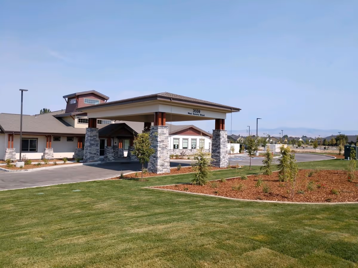 Exterior view of a senior living facility building with a covered entrance supported by stone pillars, surrounded by landscaped grass and small trees under a clear blue sky.