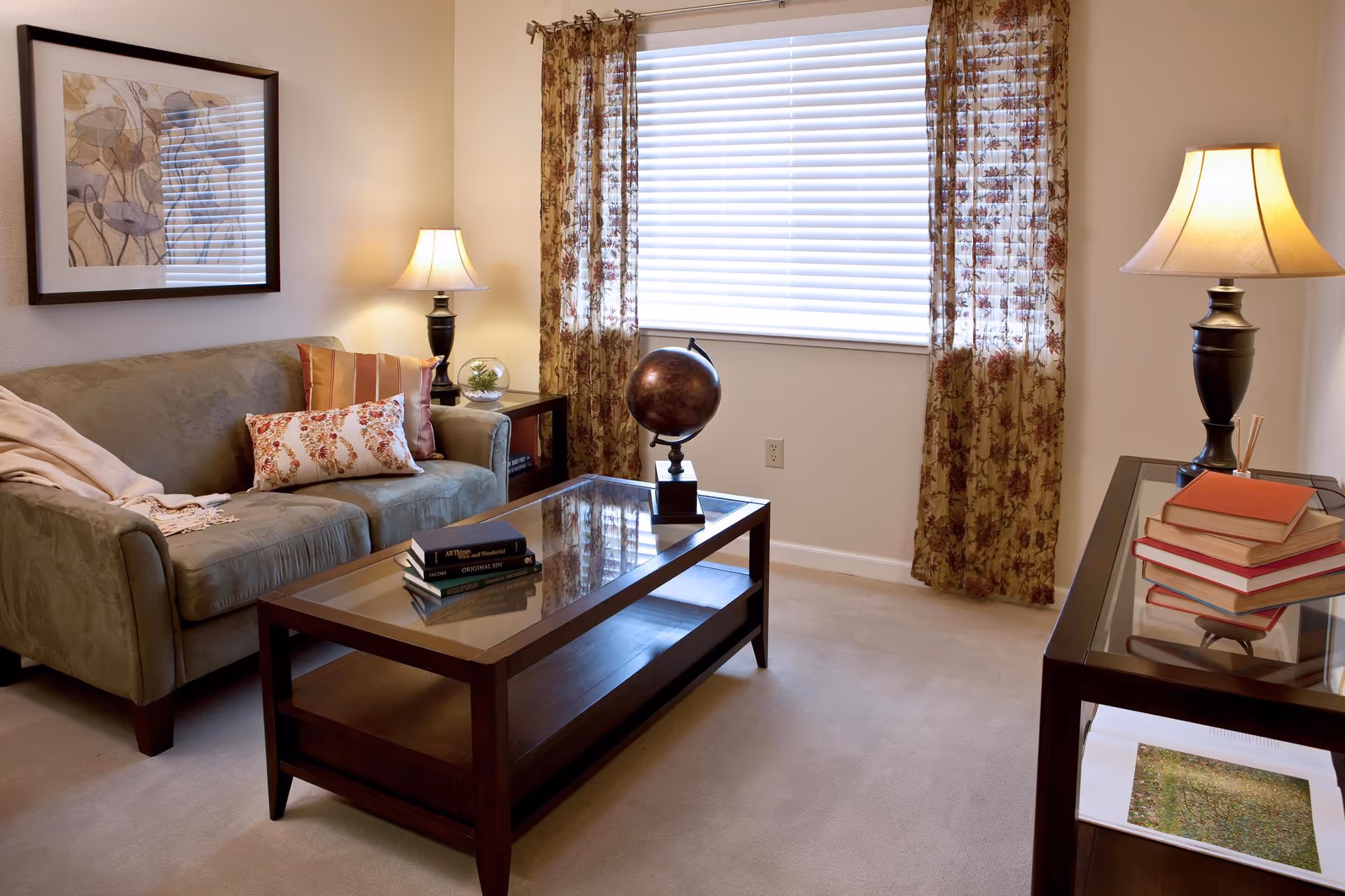 A cozy living room with a green sofa adorned with patterned pillows and a throw blanket. There is a glass-top coffee table with books and a decorative globe on it. Two table lamps with beige shades are placed on side tables, one next to the sofa and the other on a console table with more books. The room has a window with white blinds and floral curtains, and a framed floral artwork hangs on the wall above the sofa.