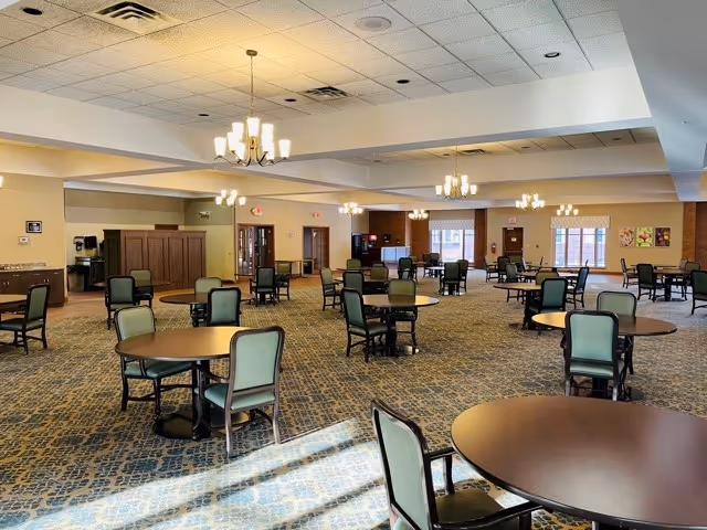 A spacious dining room with multiple round tables and green cushioned chairs arranged neatly. The room has a patterned carpet, beige walls, and several chandeliers hanging from a white ceiling with recessed lighting. Large windows allow natural light to brighten the space.