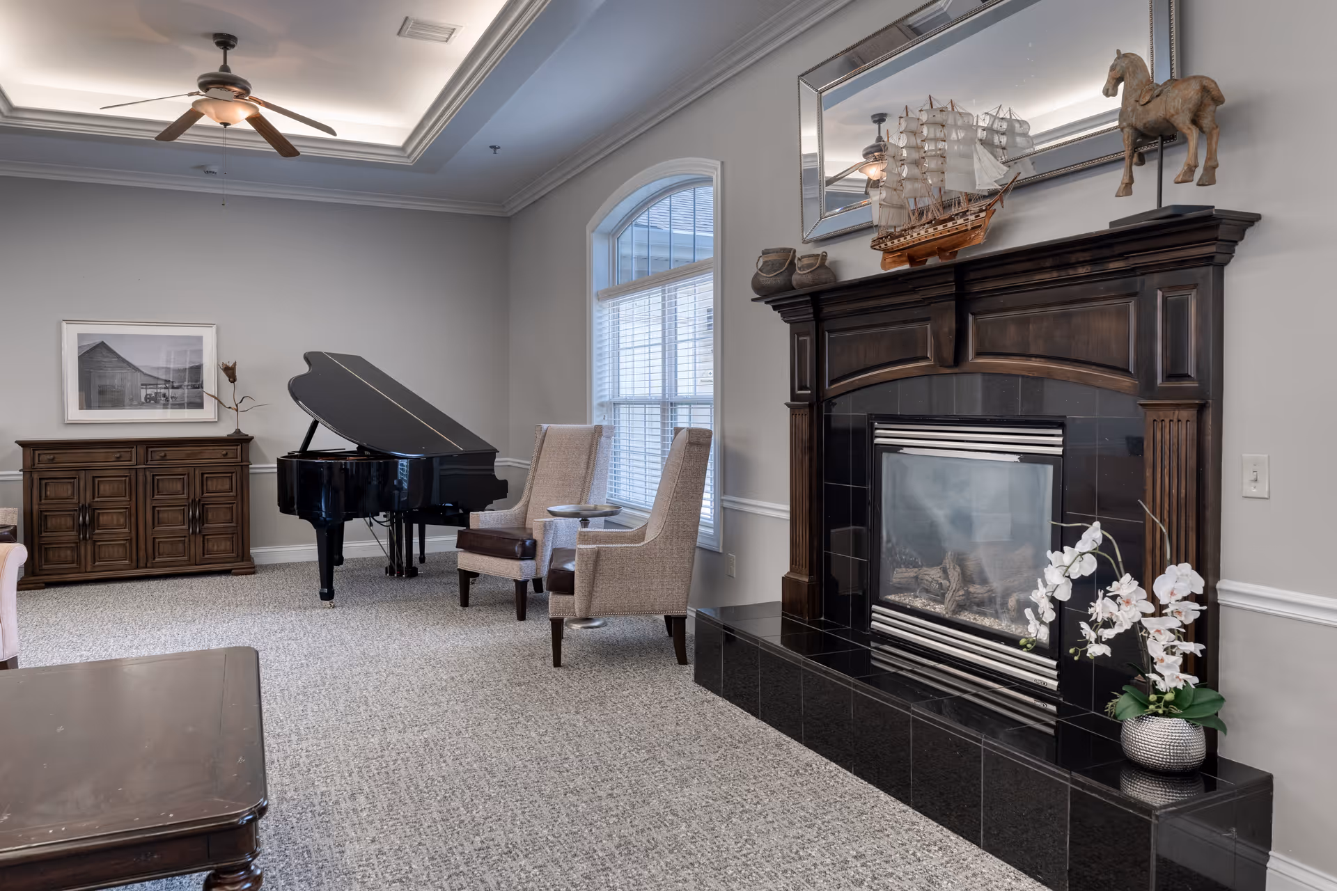 A cozy living room area featuring a black grand piano, two beige upholstered chairs with a small round table between them, a dark wooden fireplace with a decorative ship model and horse figurine on the mantel, a large mirror above the fireplace, a wooden sideboard with a framed black and white photo on the wall, and a ceiling fan with lights. The room has light gray walls, a large window with white blinds, and a carpeted floor.
